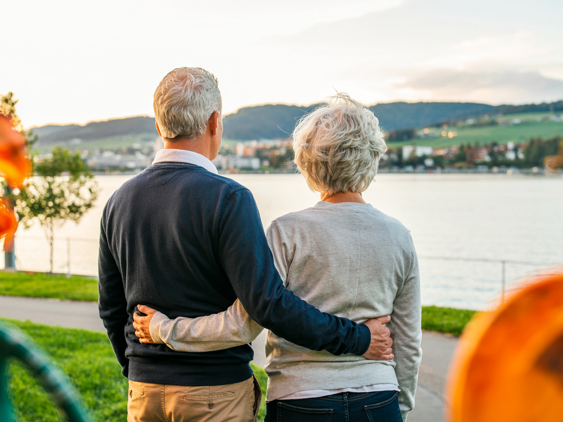 Seniorenpaar steht am Flussufer beim Sonnenuntergang und genießt gemeinsame Zeit. Illustration für Partnerschaft und Lebensqualität im Ruhestand. Konzept für AHV bei Ausgleichskasse Migros.